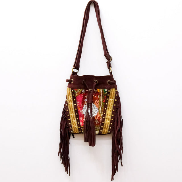 A bohemian-style bucket bag with dark brown suede-like fringe, gold beaded trim, and a colorful floral embroidered front panel, hanging against a plain white wall.