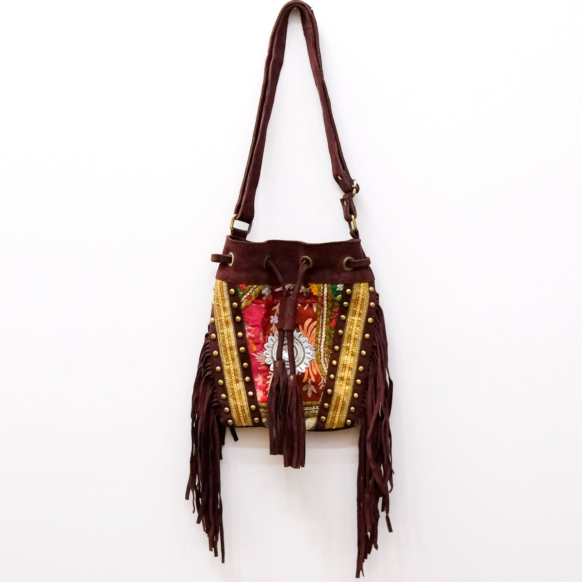 A bohemian-style bucket bag with dark brown suede-like fringe, gold beaded trim, and a colorful floral embroidered front panel, hanging against a plain white wall.